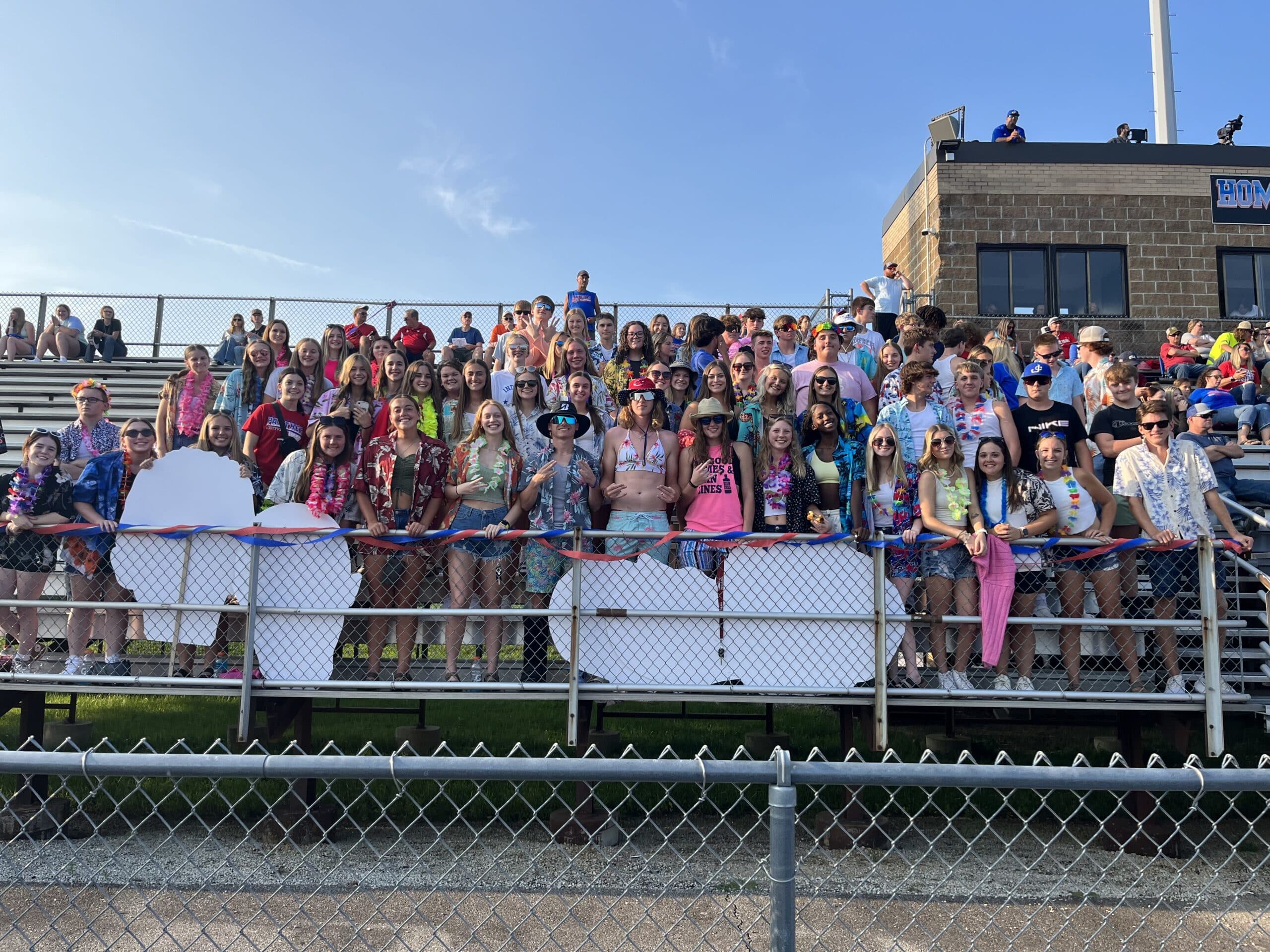 Jennings County High School student section, dressed in Hawaiian gear, standing and smiling in the stands, adding a tropical flair to the event.