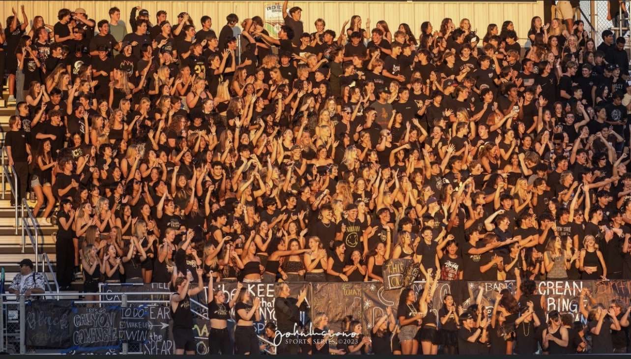 Canyon High School student section, a sea of all black attire, raise their arms in the air to cheer on their football team.