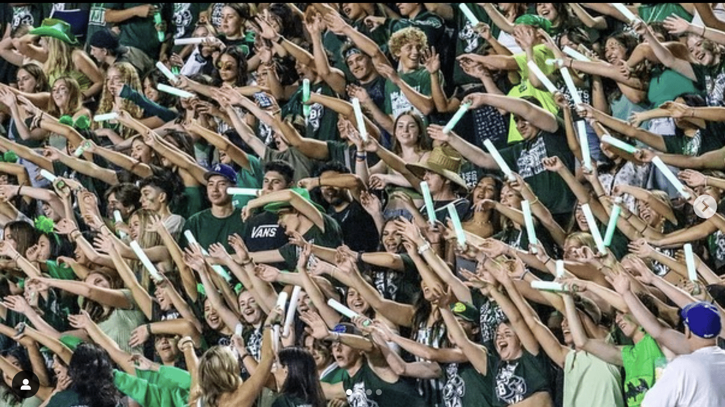 Bonita High School student section, wearing green and white school colors, enthusiastically cheers at a football game while waving glow sticks. Student leaders at the front lead the spirited cheer.