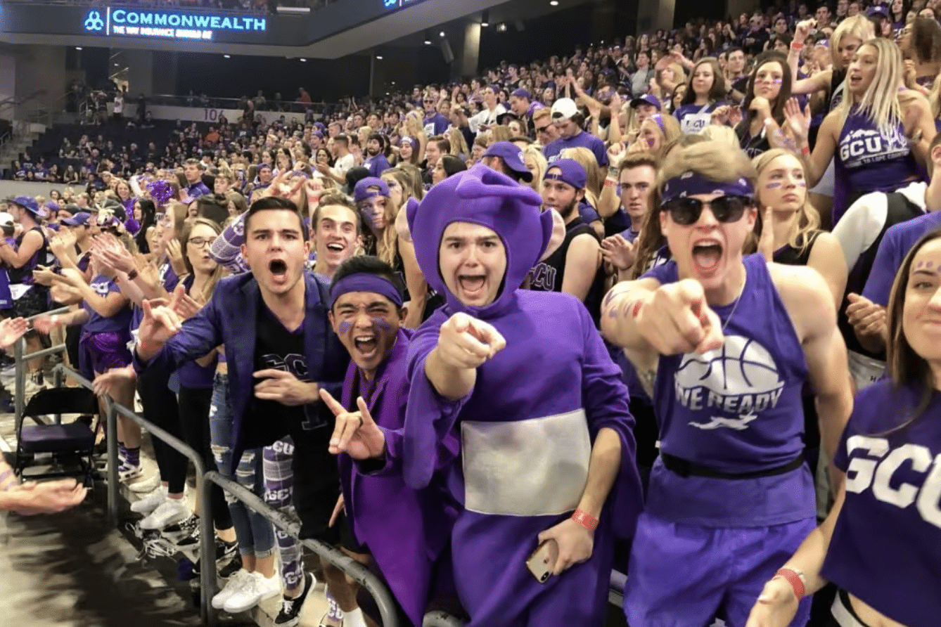 GCU student section dressed in school colors, making direct eye contact with the camera. Among them, one student stands out in a purple Teletubbie costume