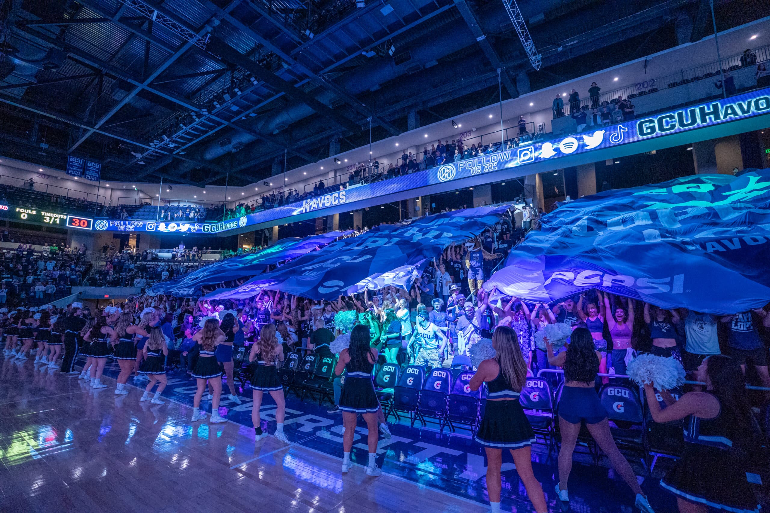 As colorful lights flash in the dark arena, the GCU Havocs student section cheers loudly and waves large banners as part of their electric pre-game atmosphere.