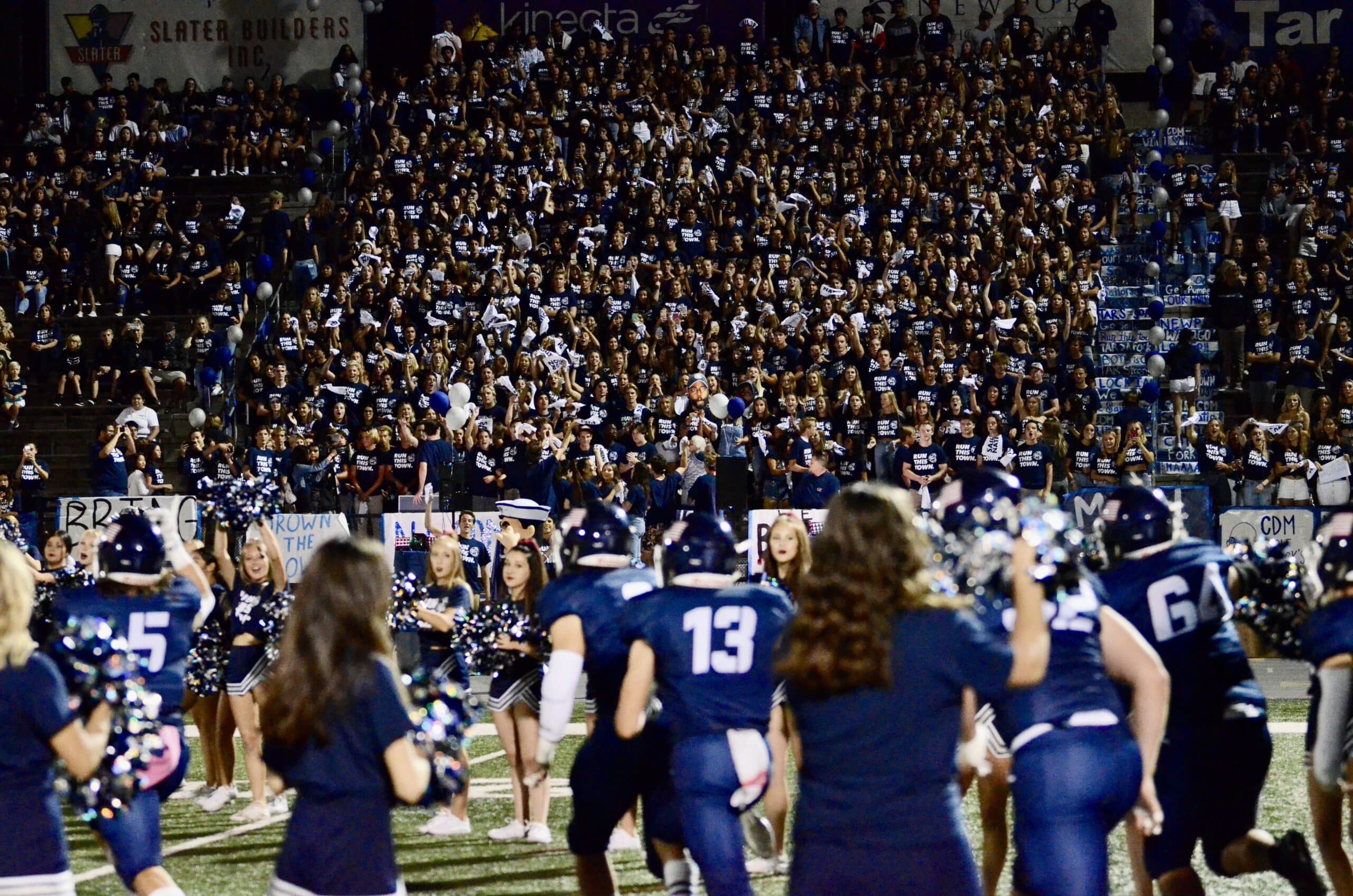 High school pep rally featuring students wearing 'run this town' merchandise, with football players and cheerleaders on the field and the spirited student section in the stands, showing their support and enthusiasm for the team.