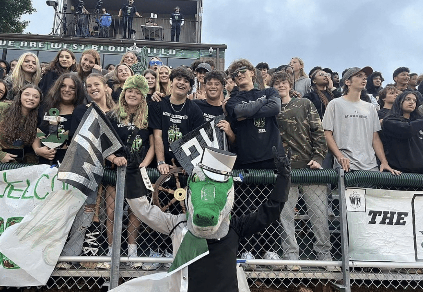 West Bloomfield High School student section smiling in the bleachers with a mascot waving their hands, creating a joyful and spirited atmosphere.